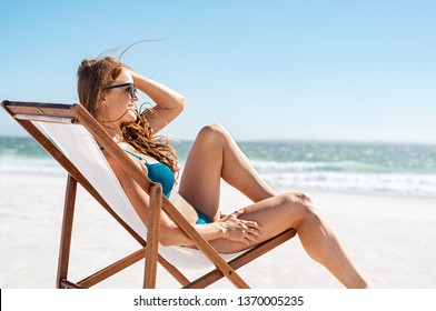 Smiling woman sunbathing on deck chair at beach. Attractive mature woman relaxing at seaside and looking the ocean. Young happy girl in bikini lying on a sun chair while wearing sunglasses.
