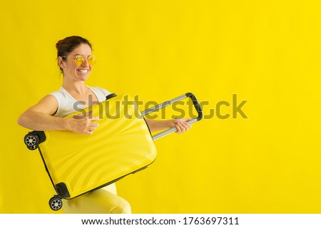 Smiling woman in sunglasses fooling around and holding a suitcase like a guitar on a yellow background. An excited girl in anticipation of a summer vacation trip simulates playing a string instrument.