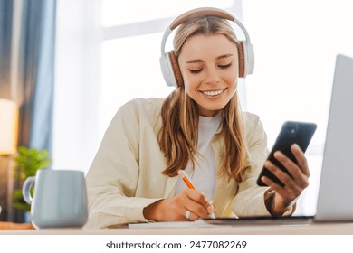 Smiling woman takes notes at her home office, wearing headphones listening music at home using mobile phone remote work. Online education concept - Powered by Shutterstock