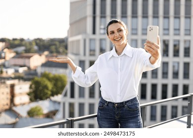 Smiling woman taking selfie on rooftop with modern buildings in background. Person enjoying outdoor city environment, capturing moment with smartphone. Casual attire, relaxed ambiance. - Powered by Shutterstock