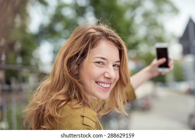 Smiling Woman Taking A Selfie On Her Mobile In An Urban Street Turning To Grin Happily At The Camera Over Her Shoulder