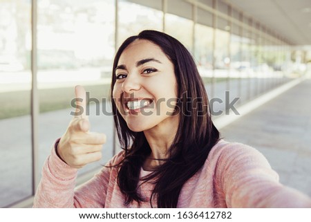 Smiling woman taking selfie photo and pointing at you outdoors. Pretty young lady looking at camera with building wall and walkway in background. Selfie and tourism concept. Front view.