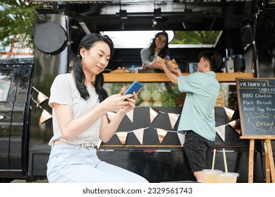 Smiling woman texting friend when boyfriend buying sandwiches in food truck - Powered by Shutterstock