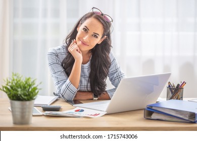 Smiling Woman With Titled Head Leaned On Her Hand Sitting At Her Office Desk.