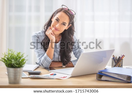Smiling woman with titled head leaned on her hand sitting at her office desk.