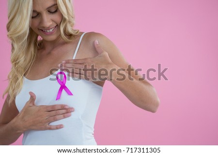 Smiling woman touching breast while looking Breast Cancer Awareness ribbon against pink background