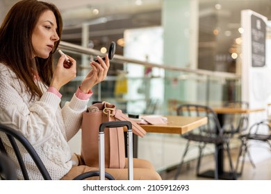 Smiling woman traveler rouge lipstick at international airport terminal before departure. Happy female doing makeup waiting flight boarding sitting at table with suitcase luggage at duty free lounge
