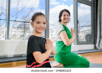 Smiling woman and tween girl sit on knees at sport mats with namaste gesture, healthy family lifestyle. Daughter and mother are looking at the camera and smiling.