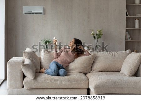 Smiling woman using air conditioner remote controller, relaxing on comfortable couch in living room alone, positive beautiful young female switching temperature on climate control system at home