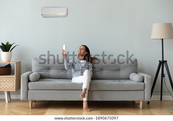Smiling woman using air conditioner remote\
controller, sitting on cozy couch at home full length, happy young\
female relaxing setting comfort temperature in modern living room,\
enjoy fresh air