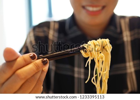Smiling woman using black plastic chopsticks to eat stir fried noodles with shrimp, ham, mushroom, vegetables and dried chili on blurred background of her boby in dark plaid shirt. (selective focus)