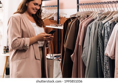 Smiling Woman Using Cell Phone While Standing At A Rack In A Clothing Store. Plus Size Female Holding Smartphone While Shopping.