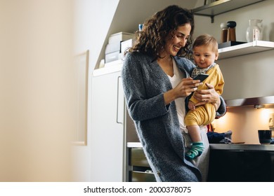 Smiling Woman Using Mobile Phone While Carrying Baby. Mother Holding Son And Texting On Cell Phone At Home.