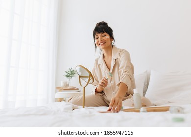 Smiling Woman Using Roller To Massage Face Sitting On Bed At Home. Woman Sitting In Bedroom Applying Cosmetics And Skin Care Products.