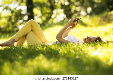 Smiling woman  using a smart phone and relaxing at the park on grass.