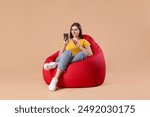 Smiling woman using smartphone on red bean bag chair against beige background