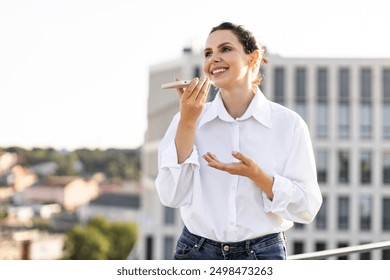 Smiling woman using smartphone voice assistant on rooftop terrace with urban background. Woman dressed in casual white shirt and jeans, enjoying daytime communication. - Powered by Shutterstock