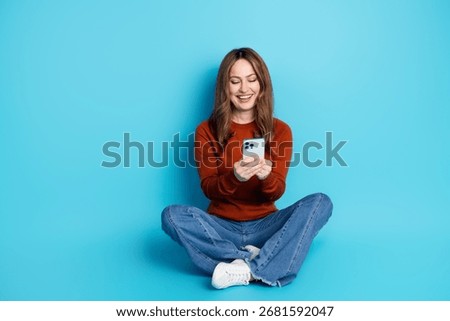 Smiling woman using a smartphone while sitting cross-legged against a blue background, wearing a casual outfit with modern style