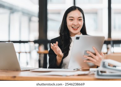 Smiling woman using tablet in modern office, engaging in video call, showcasing excitement and professionalism. Bright workspace with laptop and documents. - Powered by Shutterstock