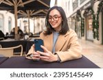 Smiling woman wear glasses sits in outdoor cafe takes selfies by smartphone. Confident woman sits in cafe dressed in coat in fall looking at phone.
