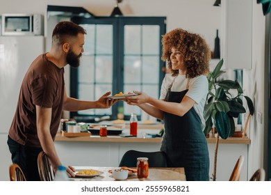 Smiling woman wearing an apron passes a plate of scrambled eggs to her partner in a brightly lit kitchen, creating a heartwarming scene of domesticity and shared meal preparation - Powered by Shutterstock