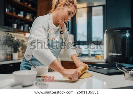 Smiling woman wearing apron using rolling pin, flattening dough on kitchen counter, preparing to make cookies, baking tray and cookie cutters nearby