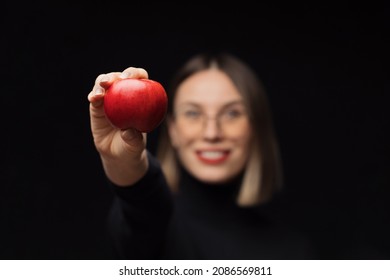 A smiling woman wearing glasses shows a red apple to camera, with red lips, over a black background. Focus on fruit