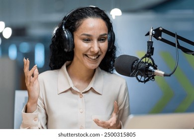 Smiling woman wearing headphones and using microphone recording podcast episode. Content creator speaking into microphone, engaging with audience in studio. Concept of podcasting, communication - Powered by Shutterstock