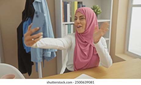 Smiling woman wearing hijab taking selfie indoors with smartphone in a bright room - Powered by Shutterstock