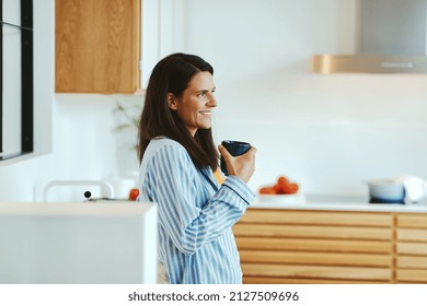Smiling woman wearing pajamas standing in her kitchen and drinking a fresh cup of coffee in the morning - Shutterstock ID 2127509696