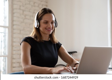 Smiling woman wearing wireless headphones working typing on notebook sit at desk in office workplace. Enjoy e-learning process, easy comfortable application usage, listen music during workday concept - Powered by Shutterstock