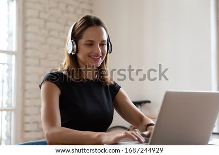 Smiling woman wearing wireless headphones working typing on notebook sit at desk in office workplace. Enjoy e-learning process, easy comfortable application usage, listen music during workday concept