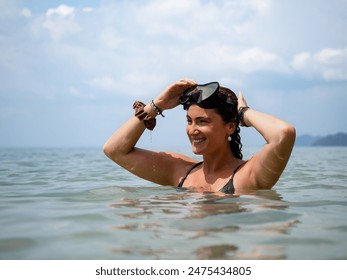 A smiling woman with wet hair adjusts her snorkeling mask while standing in the clear ocean waters. The sunny day and scenic seascape enhance her joyful and adventurous spirit - Powered by Shutterstock