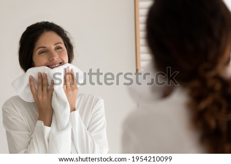 Smiling woman wiping face with white fluffy towel after shower, happy attractive young female wearing bathrobe looking in bathroom mirror, enjoying skincare beauty spa procedure, morning routine
