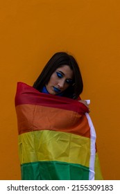Smiling Woman Wrapped Rainbow Flag While Standing Against orange background. vertical portrait