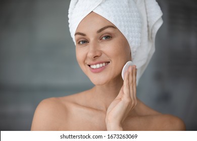 Smiling Woman With Wrapped In Towel Head Using Cotton Pad, Moisturizing Face Skin With Toner After Morning Shower. Happy Lady Looking At Camera, Doing Skincare Routine In Bathroom, Head Shot Close Up.