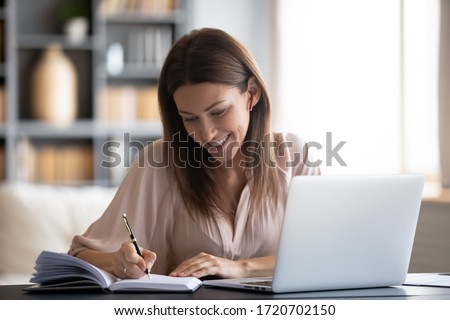 Smiling woman writing notes in notebook daily planner, planning week, working or studying at home, using laptop, beautiful female wearing wireless earphones listening to music or lecture