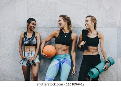Smiling women athletes in fitness clothes relaxing after workout. Three fitness women standing outdoors holding skipping rope basketball and mat after workout.