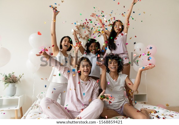 Smiling women group wear sleepwear celebrate party\
throw confetti up in air look at camera, happy girls best friends\
having fun on bed enjoy bachelorette slumber hen party with\
balloons in hotel room