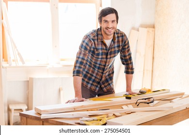 Smiling woodworker. Cheerful young male carpenter leaning at the wooden table with diverse working tools laying on it