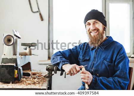 Smiling worker with a beard wearing blue jeans suit and black hat sitting on working place with wooden spoon, woodcarving instruments on table, window at background.