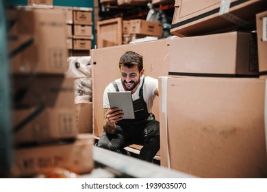 Smiling worker in overalls crouching in warehouse,using tablet for checking on inventory.