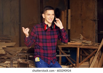 Smiling Workman Dressed In The Checkered Shirt Talking The Phone At The Sawmill. Timbers On Background.