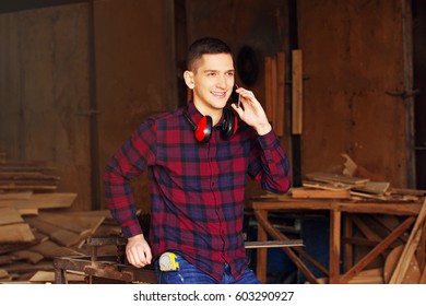 Smiling Workman Dressed In The Checkered Shirt Talking The Phone At The Sawmill. Timbers On Background.