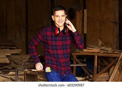 Smiling Workman Dressed In The Checkered Shirt Talking The Phone At The Sawmill. Timbers On Background.