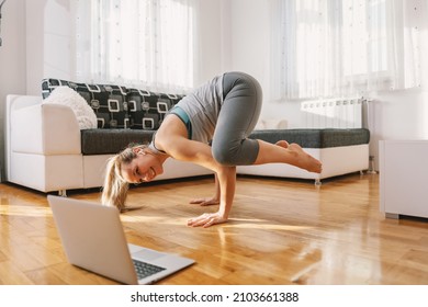 Smiling yoga instructor in crow yoga pose having online class over laptop. Home interior.