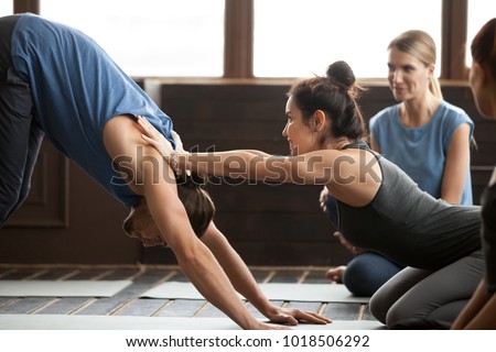 Smiling yoga teacher or pilates instructor helping young man to stretch muscles holding hands on his shoulders, sporty fit guy doing downward facing dog exercise with trainer at group class training
