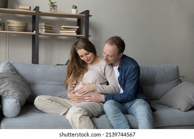 Smiling young 35s man stroking belly of pregnant wife, expressing sincere loving feelings to unborn baby, expecting childbirth together, resting on comfortable couch at home, happy parenthood concept.