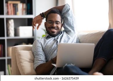 Smiling Young Adult African American Guy Using Laptop Relaxing On Sofa, Happy Millennial Single Man Lounge On Couch Holding Computer Device Watching Movies Enjoying Surfing Internet On Weekend Alone