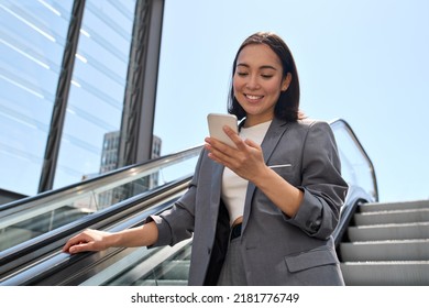 Smiling young adult Asian business woman wearing suit standing on urban escalator using applications on cell phone gadget, reading news on smartphone, browsing fast mobile internet outdoors. - Shutterstock ID 2181776749